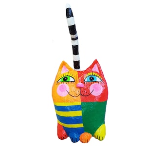 Eine handbemalte Katzenfigur. Das Gesicht der Katze ist in orange und rote Hälften geteilt, mit grünen und blauen Augen, rosa Wangen und einer rosa Nase. Der Körper ist blau und gelb gestreift, mit einem grünen unteren Teil. Der Schwanz ist schwarz-weiß gestreift.