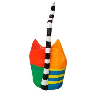 Rückansicht einer bunt bemalten Katzenfigur aus Pappmaché mit orangefarbenem und gelbem Kopfbereich, grün-blau gestreiftem unteren Körper und schwarz-weiß gestreiftem aufgerichtetem Schwanz vor hellem Hintergrund. Fotografiert frontal von hinten aus leicht erhöhter Blickrichtung.