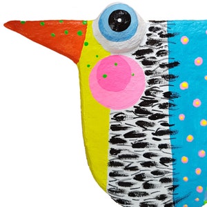 Detailreiche Nahaufnahme des farbenfrohen Gesichts einer handgefertigten Vogel Figur aus Pappmaché. Im Fokus stehen das markante, hervorstehende hellblaue Auge, der gepunktete orange Schnabel und die rosa Wange auf gelbem Grund. Das Foto zeigt die feine Textur der moderne naive Kunst Skulptur sowie die aufwendige Handbemalung mit dem blau-weißen Federmuster und den bunten Punkten. Dieser Fantasievogel als Unikat ist ein faszinierender Blickfang.