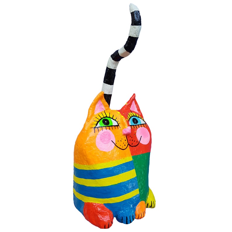 Eine skurrile, farbenfrohe Katzenskulptur. Die Katze ist in leuchtenden Farben wie Orange, Gelb, Blau, Grün und Rot bemalt. Sie hat einen gestreiften Schwanz und einen fröhlichen Gesichtsausdruck. Der Körper der Katze ist mit horizontalen blau-gelben Streifen verziert.