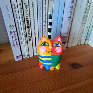 Handgefertigte Katzenfigur aus Pappmaché mit orangefarbenem Kopf, spitzen Ohren, großen grünen Augen, schwarzen Wimpern und runden rosa Wangen. Der Körper ist in Gelb, Blau, Grün und Rot bemalt und teilweise gestreift. Der lange, schwarz-weiß geringelte Schwanz ragt senkrecht nach oben. Die bunte Katze sitzt auf einem Holzregal vor einer Reihe von Büchern.