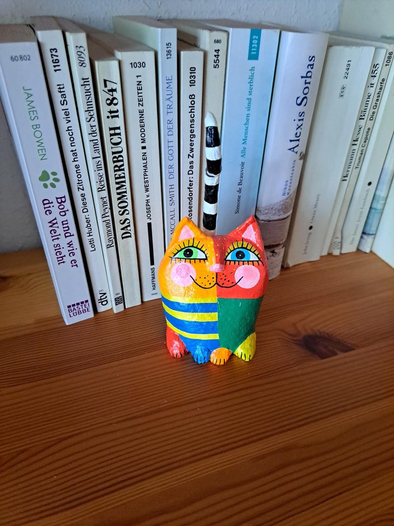 Handgefertigte Katzenfigur aus Pappmaché mit orangefarbenem Kopf, spitzen Ohren, großen grünen Augen, schwarzen Wimpern und runden rosa Wangen. Der Körper ist in Gelb, Blau, Grün und Rot bemalt und teilweise gestreift. Der lange, schwarz-weiß geringelte Schwanz ragt senkrecht nach oben. Die bunte Katze sitzt auf einem Holzregal vor einer Reihe von Büchern.