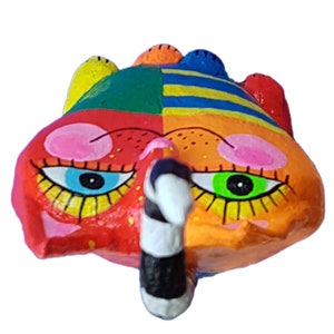 Eine farbenfrohe, abstrakte Katzenfigurine von oben fotografiert. Das Gesicht ist in Abschnitte aus Rot, Orange, Grün und Blau unterteilt. Es hat große Augen mit blauen und grünen Iris, rosa Wangen und einen schwarz-weißen schnabelartigen Vorsprung.