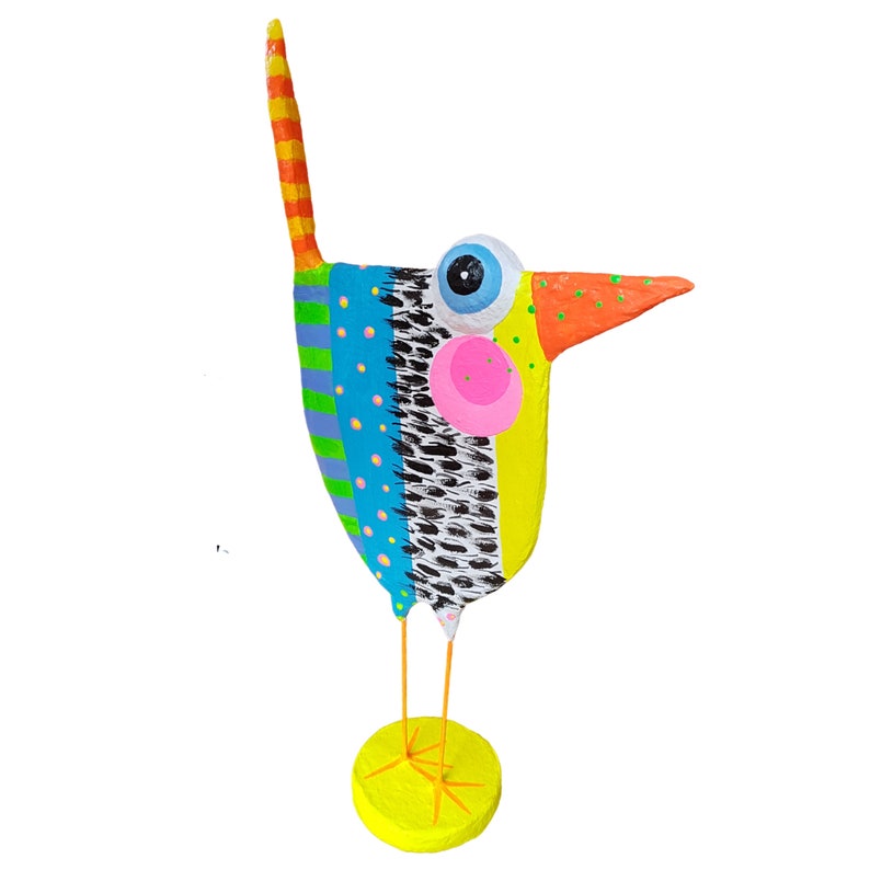 Dreiviertel-Ansicht der Vogel Figur aus Pappmaché, die das flache Profil und die räumliche Wirkung der Skulptur betont. Der Fantasievogel steht auf grazilen orangefarbenen Beinen auf einem leuchtend gelben Sockel. Sichtbar sind der gepunktete Schnabel, die markante rosa Wange auf gelbem Grund sowie das blau-schwarze Federmuster und die Punkte auf blauem Grund. Ein farbenfrohes Unikat moderner naiver Kunst als Blickfang.