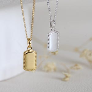 Könnte beinhalten: Zwei Anhänger-Halsketten. Eine ist goldfarben mit einem rechteckigen Anhänger und einem Perlenrand. Die andere ist silberfarben mit einem weißen rechteckigen Anhänger und einem Perlenrand. Beide hängen an filigranen Ketten.