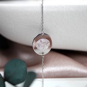 Könnte beinhalten: Ein silbernes Kettenarmband mit einem runden Anhänger, der ein detailliertes Fingerabdruckdesign zeigt.