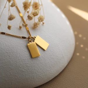 Könnte beinhalten: Eine goldfarbene Halskette mit zwei rechteckigen Anhängern. Die Kette und die Anhänger sind goldfarben. Die Halskette wird auf einer weißen Oberfläche mit getrockneten Blumen im Hintergrund präsentiert. Die Anhänger sind glatt und glänzend.