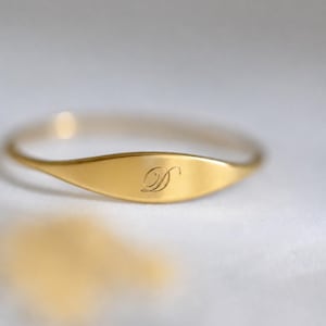 Könnte beinhalten: Ein goldfarbener Ring mit einer glatten, länglichen ovalen Platte in der Mitte. Die Platte ist mit einem einzelnen, kursiven Buchstaben D in einer feinen, fließenden Schrift graviert. Der Ring ist dünn und poliert und hat ein einheitliches Finish.