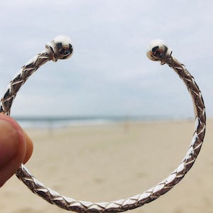 Könnte beinhalten: Ein silbernes Armreif-Armband mit geflochtenem Design und abgerundeten Enden. Das Armband wird in einer Hand mit einem verschwommenen Strandhintergrund gehalten.