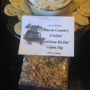 May include: A clear glass bowl filled with creamy dip, surrounded by tortilla chips on a glass plate. A packet of Cajun dip mix is in front of the bowl. The text on the label reads "Bayou Country Cookin' Louisiana Kickin' Cajun Dip".