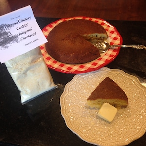 May include: A bag of "Bayou Country Cookin' Jalapeno Cornbread" mix, made in Louisiana. A red and white checkered plate with a round cornbread loaf, a slice of cornbread on a white plate with a pat of butter, and a silver serving spoon.