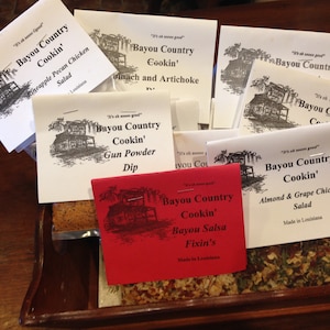 May include: A wooden tray filled with small bags of different flavored seasonings. The bags are labeled "Bayou Country Cookin'" and feature various flavors, including Gun Powder Dip, Bayou Salsa Fixin's, Pineapple Pecan Chicken Salad, Almond & Grape Chicken Salad, and Spinach and Artichoke.