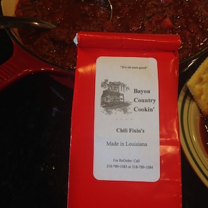 May include: Red and white bag with the text "It's oh sooo good" and "Bayou Country Cookin' Chili Fixin's Made in Louisiana" with phone numbers for reorders.