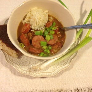 May include: A white bowl filled with a brown stew with sausage, rice, and green onions. The bowl is on a white plate with a green onion on the side.
