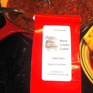 May include: A red package of "Bayou Country Cookin' Chili Fixin's" sits in front of a pot of chili. The package has a white label with text and an illustration of a building. The chili is in a red pot with a ladle.