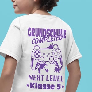 May include: White t-shirt featuring a purple video game controller graphic and text: "GRUNDSCHULE COMPLETED, NEXT LEVEL, Klasse 5". The design is printed on the back of the shirt.