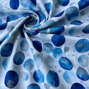 Könnte beinhalten: Weißer Stoff mit einem Muster aus blauen Aquarellformen und silbernen Glitzerpunkten. Die blauen Formen variieren in Größe und Farbton und erzeugen ein dynamisches Design. Der Stoff wird wahrscheinlich für Kleidung oder Heimdekoration verwendet.