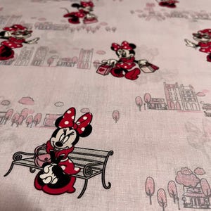 Peut inclure: Tissu rose clair avec un motif répété de Minnie Mouse en tenues à pois rouges et blancs. Minnie est représentée dans différentes poses, notamment assise sur un banc et portant des sacs à provisions. L'arrière-plan comprend des paysages urbains et des arbres stylisés.