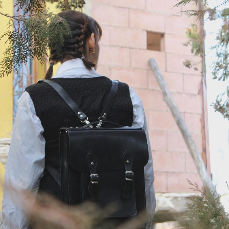 May include: Black leather backpack with two buckle closures and a top handle. The backpack has adjustable straps and is worn over a black vest and white shirt. The background includes a brick wall and foliage.