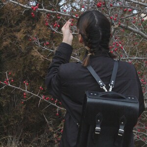 May include: A black leather backpack with silver hardware is worn by a person in a black jacket. The backpack has a top handle and two adjustable straps. The person is reaching towards a tree with red berries.