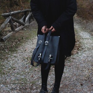 May include: Black leather backpack with silver buckles and straps. The bag has a rectangular shape with a flap closure. The straps are adjustable. The person is wearing a black coat and black leggings.