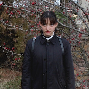 May include: A person wearing a black coat with a white collar and black leather straps. The background features a tree with red berries and a stone wall.