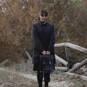 May include: A person wearing a black coat and holding a black satchel with a buckle closure. The person is standing in a natural outdoor setting with a wooden fence and brown foliage in the background.