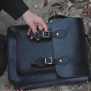 May include: Black leather satchel with a rectangular shape and a front flap. The bag features a small front pocket and two adjustable buckle closures. The bag is held by a person.