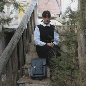 May include: A black leather satchel with two buckles and a front pocket sits on a concrete staircase. The satchel is in front of a person wearing a black dress over a white collared shirt. The person is sitting on the stairs.