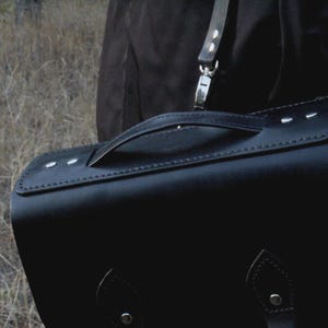 May include: A black leather satchel with a handle and a shoulder strap. The bag has a rectangular shape with rounded corners and is secured with a buckle closure. The stitching is visible along the edges, and the hardware is silver-toned.