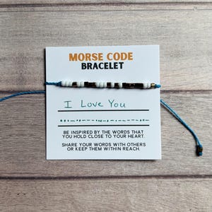 May include: A Morse code bracelet featuring alternating white and metallic dark beads on a blue cord, displayed on a white card with the text MORSE CODE BRACELET, I Love You, and inspirational phrases printed in black and orange ink.