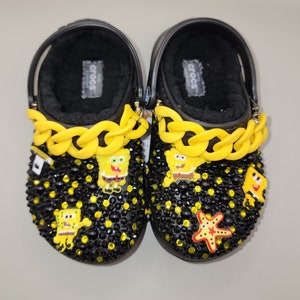 May include: Pair of black Crocs adorned with yellow chain accents, black and yellow rhinestones, and SpongeBob SquarePants character charms. The interior is lined with black faux fur, and a red starfish charm is also present.