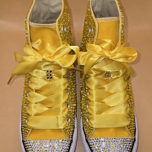 May include: A pair of yellow high-top sneakers embellished with sparkling rhinestones. The toe caps are covered in silver and yellow gems. The shoes have yellow satin ribbon laces and white rubber soles.
