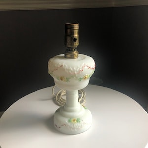 May include: White glass table lamp with a brass socket. The lamp features a decorative floral design with red, yellow, and green accents. The lamp has a round base and a long stem. The electrical cord is visible.