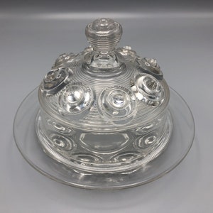 May include: Clear glass butter dish with a domed lid and a circular base. The lid features a central knob and decorative circular accents. The dish is designed to hold butter and is made of transparent glass.