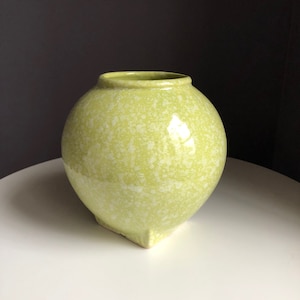 May include: A round, chartreuse-colored ceramic vase with a speckled white pattern. The vase has a wide, open top and a slightly flattened base with three small feet. The vase is a decorative item.