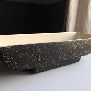 May include: A rectangular ceramic planter with a black exterior and cream-colored interior. The black surface is adorned with gold abstract lines. The planter is approximately 14 inches long, 6 inches wide, and 4 inches tall.