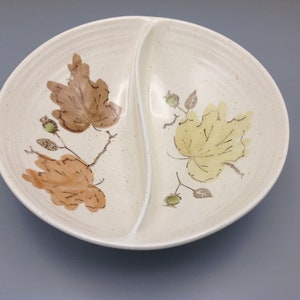 May include: Cream-colored, divided serving dish with leaf designs. The dish features a central divider and is decorated with brown and yellow leaves and green berries. The dish has a speckled texture.