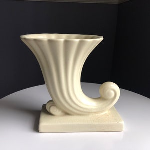 May include: Cream-colored ceramic cornucopia vase with a square base and a curved, horn-like shape. The vase has a fluted design and a decorative scroll detail. A vintage decorative piece.