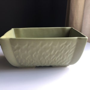 May include: A square, light green ceramic planter with a textured, woven pattern on the sides. The planter has a slightly flared rim and sits on a white surface. The background is a mix of black and neutral tones.