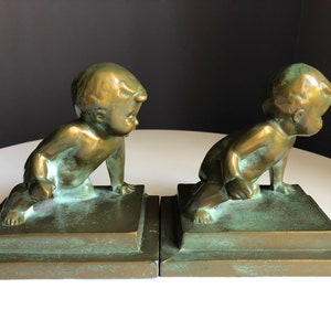 May include: A pair of bronze bookends, each featuring a crawling baby figure. The figures are positioned on square bases, with a weathered green patina. The bookends are designed to stand upright and support books.