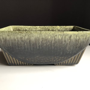 May include: A rectangular ceramic planter with a gradient glaze, transitioning from light green to dark gray. The planter has a curved lower section with vertical lines and measures approximately 12 inches long, 6 inches wide, and 4 inches tall.