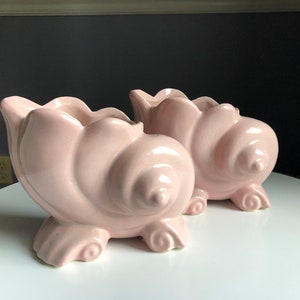May include: Two light pink ceramic seashell planters. Each planter has a spiral design and rests on three small feet. The top edge of each planter has a scalloped design. These decorative planters are ideal for succulents or small plants.