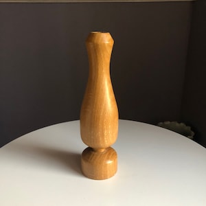 May include: A light brown wooden vase with a rounded base and a slender, elongated body. The vase has a smooth, polished finish and a small opening at the top. The vase is displayed on a white surface.