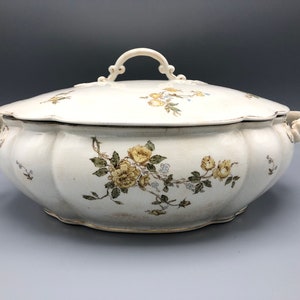 May include: Cream-colored, lidded serving dish with a floral design. The oval-shaped dish features a scalloped edge and a handle on the lid. Yellow and green floral patterns adorn the dish, with additional handles on either side. A classic piece for serving.