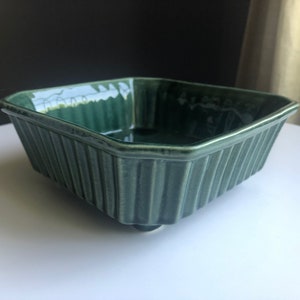 May include: A square, dark teal ceramic baking dish with vertical ribbed sides. The dish has a slightly raised base and a glossy finish. The interior is smooth and reflects light. This vintage-style dish is suitable for baking and serving.