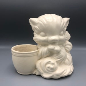 May include: A cream-colored ceramic cat planter. The cat is sitting with a small pot attached to its side. The cat has a cute, cartoonish design with large eyes and a fluffy appearance. The planter is suitable for small plants or flowers.
