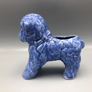 May include: A blue ceramic poodle planter with a textured surface. The poodle is standing on four legs with a small opening on its back for plants or flowers. The planter is a light blue color.
