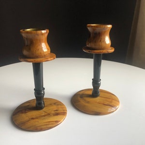 May include: A pair of wooden candle holders with black painted stems and a brass top. The holders have a wood grain pattern and are on a white surface.