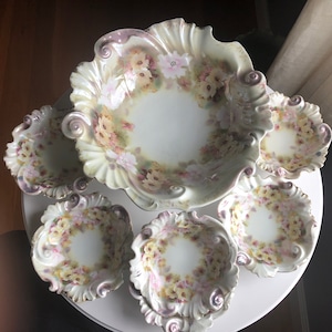 May include: A set of antique porcelain serving dishes. The set includes a large, scalloped bowl and several smaller bowls. Each piece is decorated with a floral pattern in shades of pink, yellow, and white, with a pearlescent finish and a decorative rim.